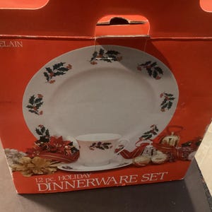 May include: A red box displaying a 12-piece holiday porcelain dinnerware set. The set includes a plate, cup, and saucer, all decorated with holly and berry designs. The box has a handle and the text "12 pc. HOLIDAY DINNERWARE SET".