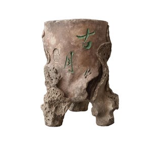 May include: A weathered, brown ceramic planter with a textured, rock-like appearance. The planter features green Chinese characters and a circular seal. The planter has three sturdy legs and is suitable for indoor or outdoor use.