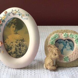 May include: Two oval-shaped porcelain picture frames with floral embellishments. The larger frame has a vintage photo of a young girl in a white dress. The smaller frame has a photo of two children in a heart-shaped opening and a small brown rabbit figurine.