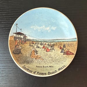 May include: A round ceramic souvenir plate featuring a vintage illustration of a crowded beach scene at Revere Beach, Massachusetts. The image depicts people in period clothing on the sand under a blue sky, with the text Souvenir of Revere Beach, Mass.