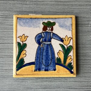 May include: A hand-painted ceramic tile featuring a woman in a blue dress and green hat standing in a field of yellow flowers. The tile has a yellow border and a blue and white background.