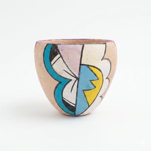 May include: A small, beige ceramic bowl with a colorful abstract design. The design features geometric shapes in blue, yellow, pink, and black. The rim of the bowl is painted purple.
