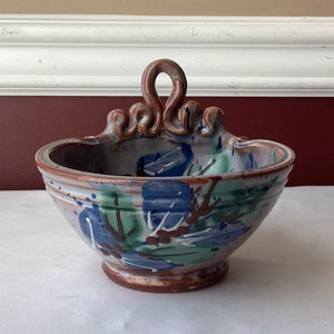 May include: A small, hand-painted ceramic bowl with a loop handle. The bowl has a brown, white, blue, and green glaze with a floral design.