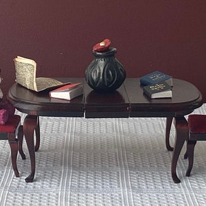May include: A dark wood table with a burgundy velvet chair and a teddy bear sitting on it. There are books and a black vase with a red lid on the table.