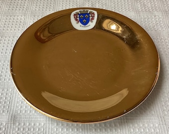 May include: A gold-colored ceramic plate with a white crest featuring a blue shield with three fleurs-de-lis and the word "Wiesbaden" underneath.