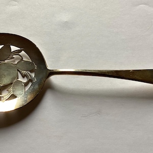 May include: A silver serving spoon with a decorative cut-out design of a fruit and leaves. The spoon has a long, slender handle.