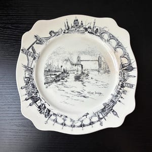 May include: A cream-colored ceramic plate with a scalloped edge, featuring a black ink illustration of the London skyline. The center depicts Tower Bridge over the River Thames, with the text Tower Bridge written below the bridge structure.