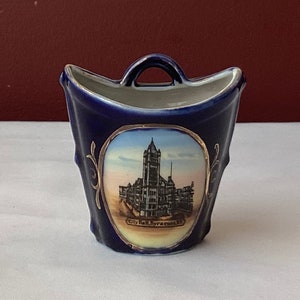 May include: A small, blue ceramic souvenir cup with a gold trim and a picture of the City Hall in Syracuse, NY.