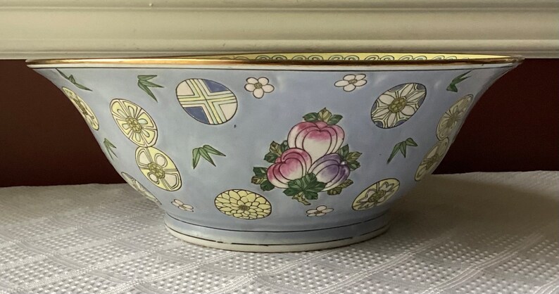 May include: A large, light blue porcelain bowl with a gold rim. The bowl is decorated with a floral pattern in shades of pink, yellow, and green. The bowl is perfect for serving fruit, salads, or other dishes.