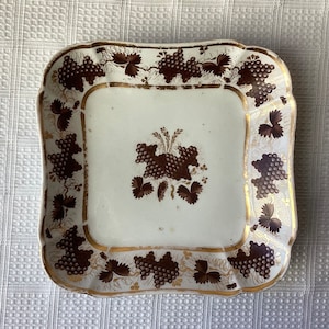 May include: A white square ceramic plate with a gold rim and brown grapevine design. The plate has a scalloped edge and is decorated with brown grapes and leaves.