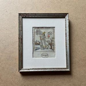 May include: Framed artwork featuring a detailed illustration of a figure holding objects, possibly a religious or historical scene. The artwork is in a silver-toned frame with a white mat. The illustration is in muted tones with a signature at the bottom.
