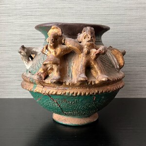 May include: An antique, hand-painted ceramic vase with a green base, brown rim, and gold accents. The vase features relief sculptures of two figures. The vase has visible cracks, adding to its aged appearance.