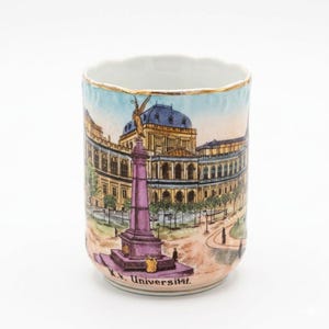 May include: A cylindrical porcelain mug featuring a hand-painted illustration of a large building and a tall purple monument. The text K.K. Universität is printed at the base. The rim is finished with a thin gold-colored trim against a white background.
