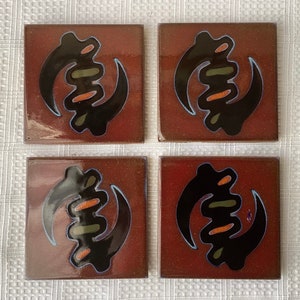 May include: Set of four brown ceramic tiles with a black and orange Adinkra symbol. The symbol is a stylized representation of a hand holding a staff.