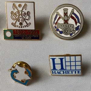 Op de afbeelding: Vier vintage emaille-speldjes. De eerste speld is wit met een gouden rand en heeft twee gekruiste polosticks met de tekst "Polo de Paris" en "1892-1992 Centenaire du Polo de Paris". De tweede speld is grijs met een gouden rand en heeft een kanon met een rood-wit-blauwe vlag. De derde speld is wit met een gouden rand en heeft een gestileerde hartvorm. De vierde speld is wit met een gouden rand en heeft een blauwe "H" met de tekst "Hachette".