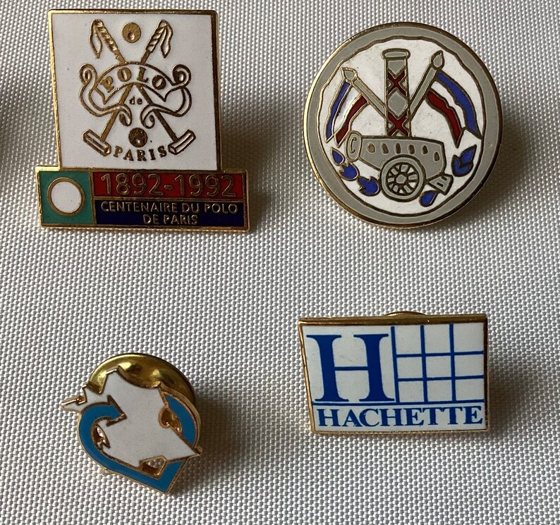 Op de afbeelding: Vier vintage emaille-speldjes. De eerste speld is wit met een gouden rand en heeft twee gekruiste polosticks met de tekst "Polo de Paris" en "1892-1992 Centenaire du Polo de Paris". De tweede speld is grijs met een gouden rand en heeft een kanon met een rood-wit-blauwe vlag. De derde speld is wit met een gouden rand en heeft een gestileerde hartvorm. De vierde speld is wit met een gouden rand en heeft een blauwe "H" met de tekst "Hachette".