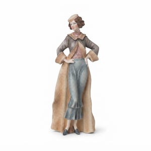 May include: A porcelain figurine of a person wearing a tan coat with fur-like trim, a pink top, and a blue skirt. The figure wears a small tan hat and dark shoes, standing with hands on hips. The sculpture features a matte finish and muted colors.