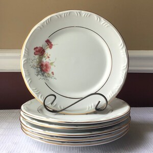 May include: A stack of five white dinner plates with a gold rim and a floral design. The plates have a raised edge and a slightly scalloped rim.