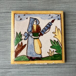May include: A hand-painted ceramic tile featuring a woman in a blue dress and yellow skirt holding a long, blue and white object. The tile has a yellow border and is decorated with green and brown foliage.