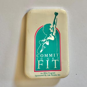 May include: A rectangular white pinback button featuring a teal arch graphic with a silhouette of a person lifting a dumbbell overhead. The text reads COMMIT TO GET FIT, An IRSA Program, Sponsored By Life Fitness, Inc. in dark green lettering.