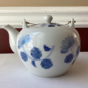 May include: White ceramic teapot with a blue floral design. The teapot has a rounded body, a curved spout, and a lid with a small knob. The handle is made of a thin metal wire. The floral pattern is repeated on the lid.