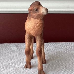 May include: A detailed figurine of a baby camel, standing on a white surface. The camel is primarily tan and brown, with a detailed face and realistic features. The figurine is likely made of ceramic or a similar material.