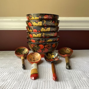 May include: A set of six black and red nesting bowls with floral and berry designs, and four wooden spoons with similar designs. The bowls are stacked on top of each other, and the spoons are laid out in front of them.