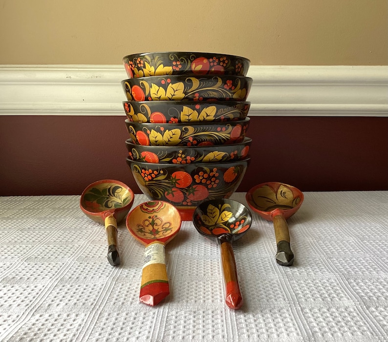 May include: A set of six black and red nesting bowls with floral and berry designs, and four wooden spoons with similar designs. The bowls are stacked on top of each other, and the spoons are laid out in front of them.
