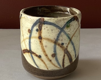May include: A small, white ceramic cup with brown, blue, and green abstract designs. The cup has a rounded bottom and a slightly flared rim.