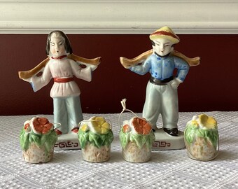 May include: Two porcelain figurines of Asian people carrying baskets on their shoulders. The figurines are painted in bright colors and have detailed facial features. The baskets are filled with flowers and leaves. The figurines are standing on a white tablecloth.