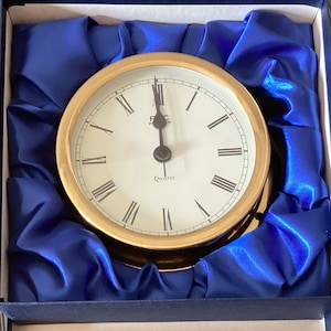 May include: A gold-toned clock with Roman numeral markings and a quartz movement. The clock is set in a blue velvet box.