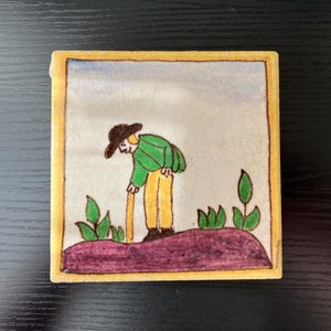 May include: A hand-painted ceramic tile with a yellow border depicting a person in green and yellow clothing holding a stick, standing on a purple hill with green plants.