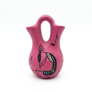 May include: A pink ceramic vase with a unique handle and a Kokopelli design. The vase features a hand-painted Kokopelli figure in black, white, and blue, along with musical notes. The vase is a decorative item.