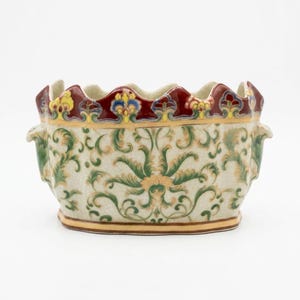May include: An oval ceramic planter featuring a cream-colored base with a fine crackle glaze finish. The exterior is decorated with intricate green and gold scrolling foliage patterns. The scalloped rim is accented with deep red and yellow floral motifs.