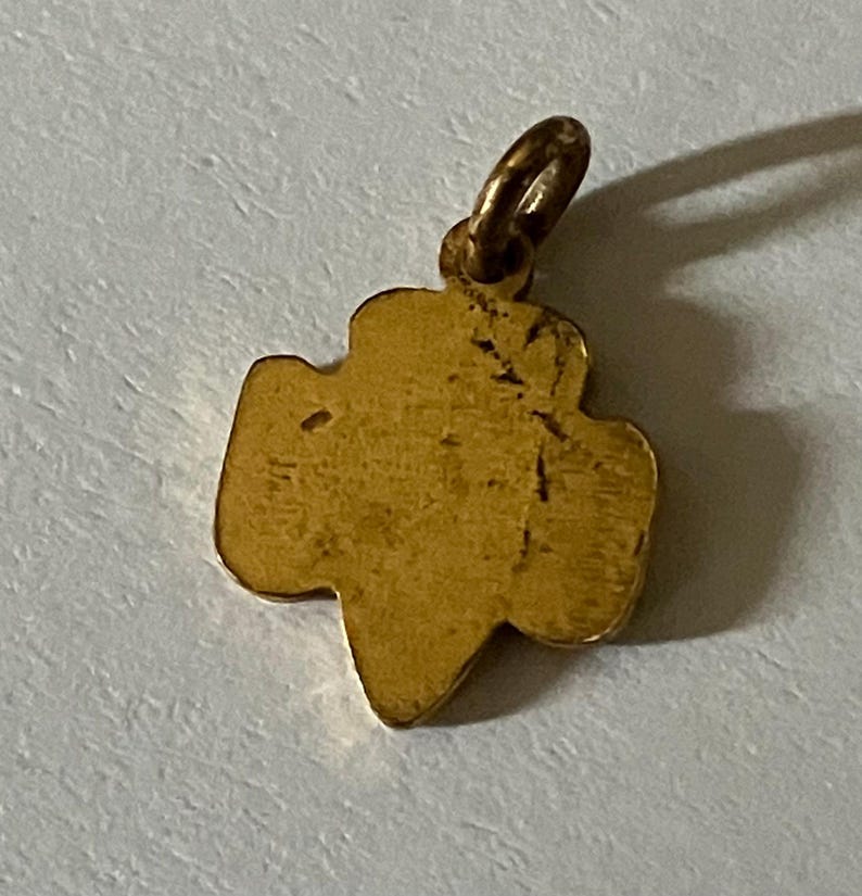 May include: A gold-colored pendant in the shape of a trefoil, with a small loop at the top for a chain. The pendant has a slightly worn appearance, suggesting it is vintage or antique. The trefoil shape is a classic symbol.