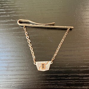 May include: Silver tie clip with a chain and a rectangular pendant with the letter 'T' in the center.