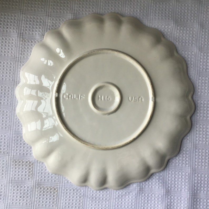 May include: White ceramic serving plate with a scalloped edge. The bottom of the plate has the words "CALIF. HI6 USA" printed on it.