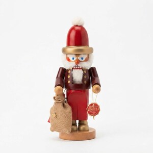 May include: A wooden nutcracker figure wearing a red hat with a white pom-pom, a dark brown jacket with gold buttons, and red trousers. It has a white beard, blue eyes, and holds a tan burlap sack. A small red tag with text is attached to its arm.