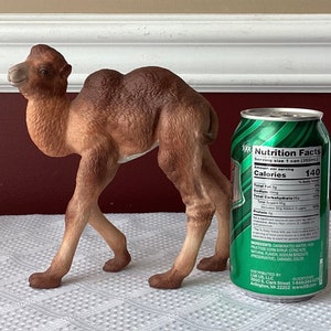 May include: A brown camel figurine stands next to a green soda can. The camel is detailed with a realistic appearance. The soda can has a green and white label with nutrition facts.