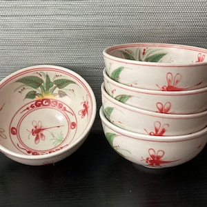 May include: A stack of six white ceramic bowls with red and green painted designs. Each bowl features a red dragonfly and floral pattern. One bowl is separate, displaying the interior design.