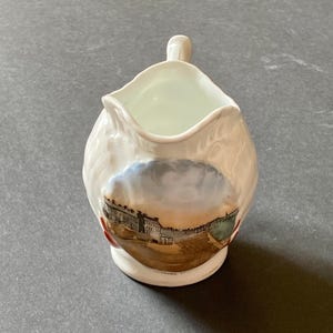 May include: A white ceramic creamer with a handle and a spout. The creamer features a scenic illustration of a building with a blue sky and brown landscape. The creamer is a decorative item.