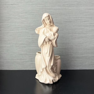 May include: A white ceramic planter shaped like a woman holding a baby. The planter has a smooth, matte finish and is a vintage style.