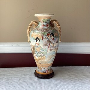 May include: A large, ceramic vase with gold accents and a floral design. The vase features two women in traditional Japanese clothing, painted in delicate detail. The vase has two handles and stands on a wooden base.