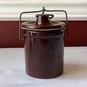 May include: A brown ceramic jar with a lid and a wire bail handle. The jar has a white rim around the top.