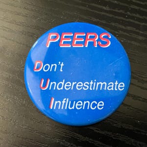May include: A round, blue button with the words "PEERS Don't Underestimate Influence" in white and red lettering. The button is a simple design, with a glossy finish.