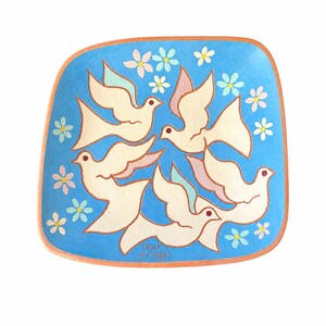May include: A square ceramic plate with rounded corners featuring a bright blue background, four cream-colored doves in flight, and small pink and light blue flowers. The text Chile is printed in the lower center. The rim is a natural terracotta color.