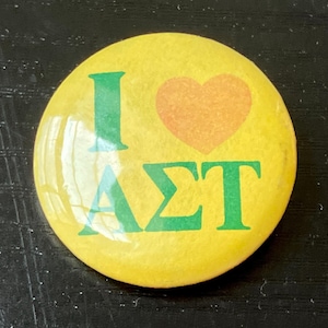 May include: A round, yellow button with the text "I" and "AST" in green, and an orange heart symbol. The button is a pinback style, likely for a sorority or organization. The button is on a dark surface.