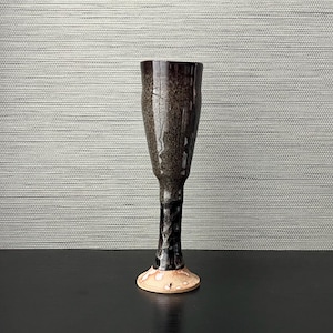 May include: A tall, handcrafted ceramic goblet with a flared top and a slender stem. The goblet features a dark brown glaze with speckled details, transitioning to black at the stem and a light tan base. The goblet stands approximately 7 inches tall.