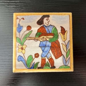 May include: A hand-painted ceramic tile featuring a man playing a lute. The tile has a white background with colorful floral designs and a yellow border.