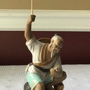 May include: A ceramic figurine of a man holding a fishing pole, sitting on a rock. The man is wearing a white shirt, blue pants, and a brown hat. The figurine is painted in shades of brown, white, and blue.